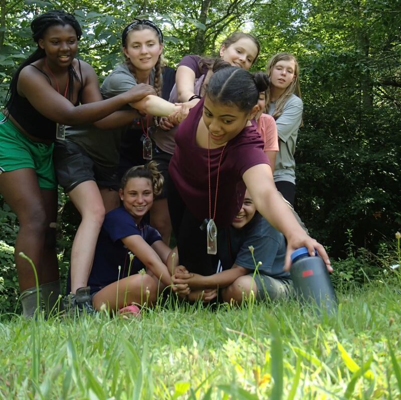 A group of young women are gathered in a grassy area, possibly a park or camp. One woman is reaching down to pick up a water bottle. The others are standing or kneeling around her, some with their arms linked or resting on each other's shoulders. They appear to be interacting in a friendly and supportive manner.